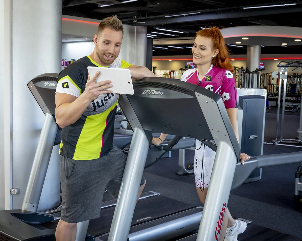 A fitness trainer guiding a woman on a treadmill using a tablet in a gym setting.