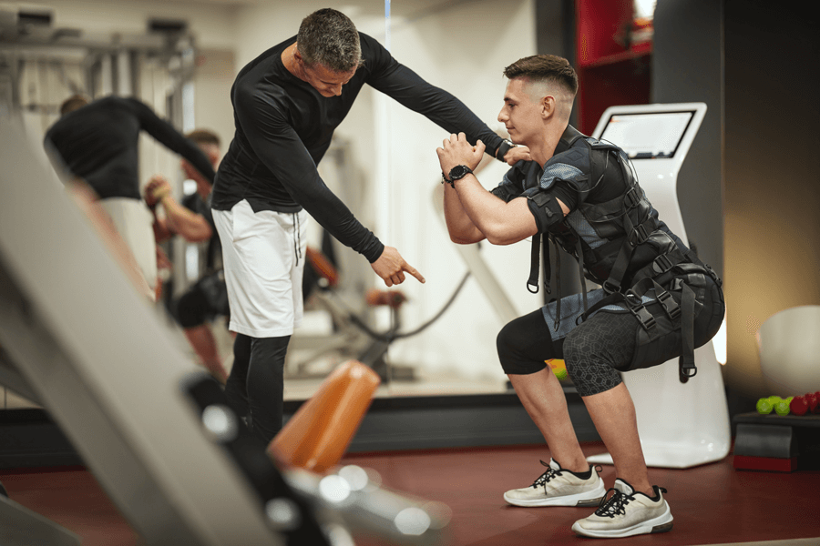 Personal trainer guiding a young man doing squats while wearing an EMS suit in a modern gym.