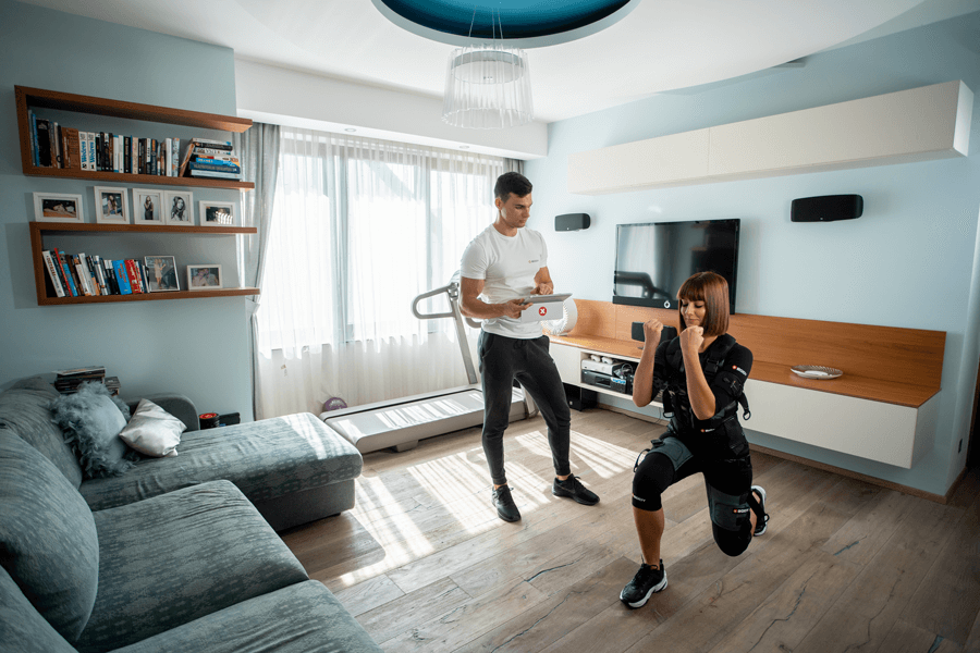 A woman performing lunges with EMS gear in a living room, assisted by a personal trainer holding a tablet.