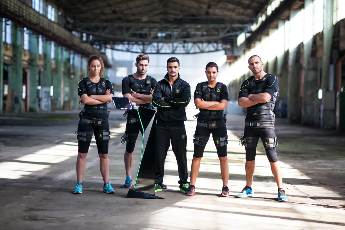 Group of five fitness trainers in EMS suits standing confidently in an industrial-style gym.